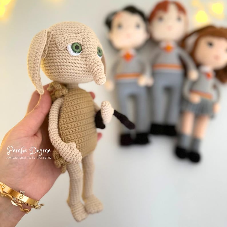 May include: A handmade crochet doll of a character with large ears, green eyes, and a long nose, holding a black wand. The doll is tan and brown. In the background are three other dolls in school uniforms. The text "Pembe Dugme AMIGURUMI TOYS PATTERN" is visible.