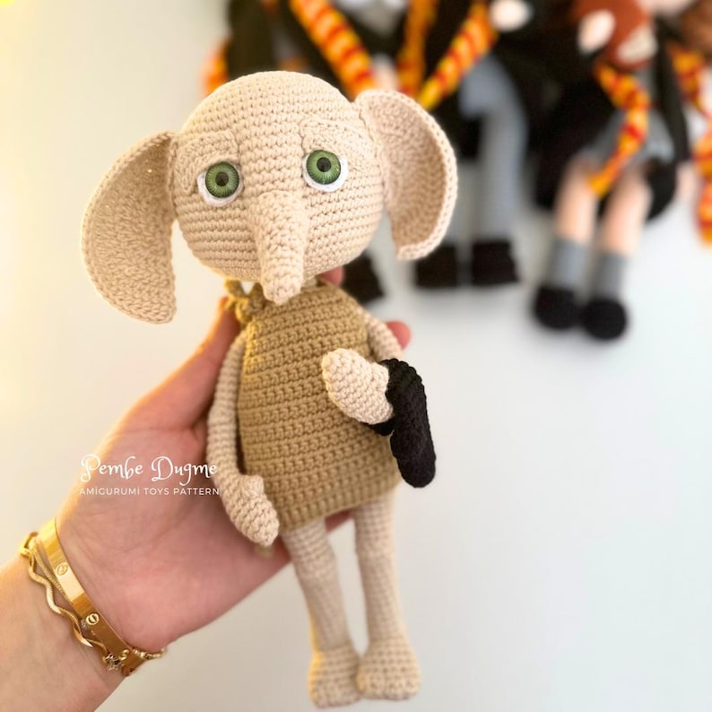 May include: A handmade crochet doll in the likeness of a character with large ears and green eyes. The doll is beige and tan, with a black item in its hand. The doll is held in a person's hand. The doll is an amigurumi toy.