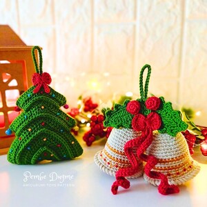 Op de afbeelding: Twee gehaakte kerstversieringen, een groene kerstboom en een paar witte bellen met rode hulst en een rood lint. De versieringen hangen aan een touwtje.
