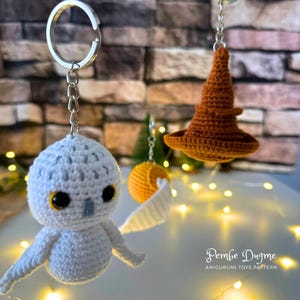 May include: A white crocheted owl keychain with a brown crocheted Sorting Hat keychain. Both keychains are hanging from silver key rings. The background is a brick wall with string lights.