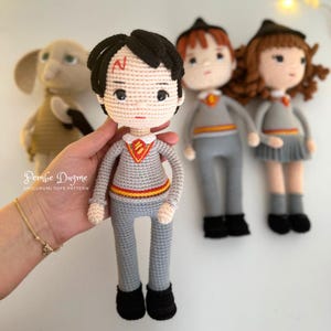 May include: Crocheted Harry Potter doll with black hair and a lightning bolt scar, wearing a gray sweater and pants. Other crocheted dolls of characters from the Harry Potter series are in the background.