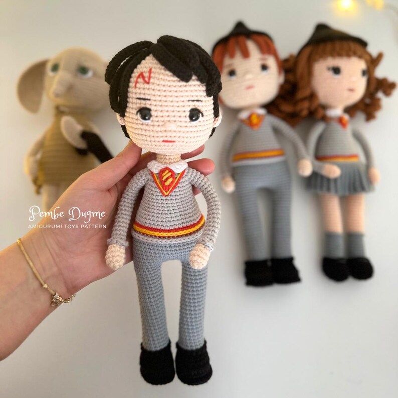 May include: Crocheted Harry Potter doll with black hair and a lightning bolt scar, wearing a gray sweater and pants. Other crocheted dolls of characters from the Harry Potter series are in the background.