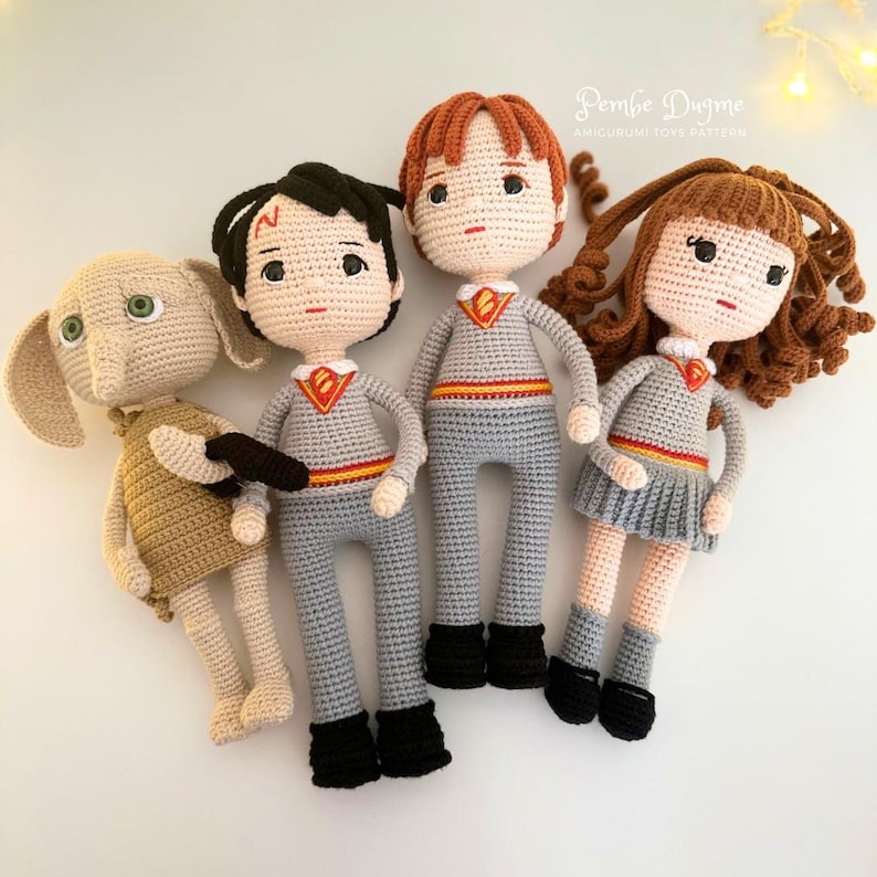May include: Four handmade crochet dolls, including Dobby, Harry Potter, Ron Weasley, and Hermione Granger. The dolls are in gray and tan outfits with black shoes. The Harry Potter dolls have a red and gold emblem on their chests. The dolls are arranged on a white surface.