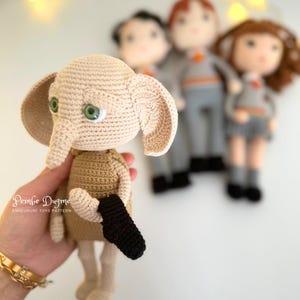 May include: A crocheted doll of a character with large ears, green eyes, and a black sock. The doll is beige and brown, with a black sock in its hand. Other crocheted dolls are in the background. The text "Pembe Dugme" is visible.