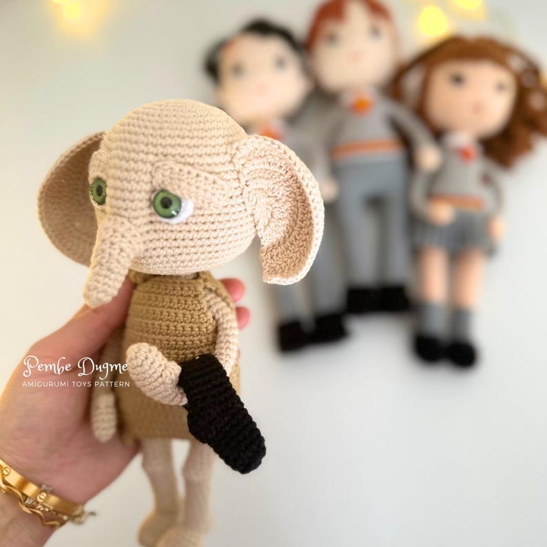 May include: A crocheted doll of a character with large ears, green eyes, and a black sock. The doll is beige and brown, with a black sock in its hand. Other crocheted dolls are in the background. The text "Pembe Dugme" is visible.