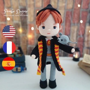 Peut inclure: Poupée en crochet amigurumi d'un jeune garçon aux cheveux roux, portant une robe noire avec une écharpe jaune et rouge, et tenant un rat en crochet gris. La poupée est debout sur une surface blanche. L'image comprend également un texte superposé qui dit "Pembe Dugme AMIGURUMI TOYS PATTERN" et trois drapeaux : américain, français et espagnol.