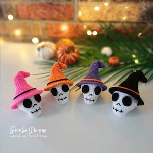 May include: Four crocheted skull ornaments with witch hats in pink, orange, purple, and black. Each skull has black eyes and a stitched mouth. The hats have contrasting trim. The background includes small pumpkins and a brick wall.