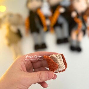 May include: A small, crocheted book with a tan interior and a rust-colored cover is held in a hand. The book is part of a collection of amigurumi toys, with several figures visible in the background. The text "Pembe Dugme" is visible.