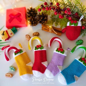 May include: Five crocheted Christmas stockings in different colours, including red, yellow, purple, blue, and green. Each stocking has a white cuff and a candy cane or mitten-shaped top. The stockings are arranged on a white surface with a small snowman and a red gift box in the background.