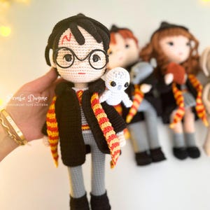 May include: Crocheted doll of a character with black hair, round glasses, and a lightning bolt scar. The doll wears a black robe, gray pants, and a red and gold scarf. A small white owl sits on the doll's shoulder. Other dolls are in the background.