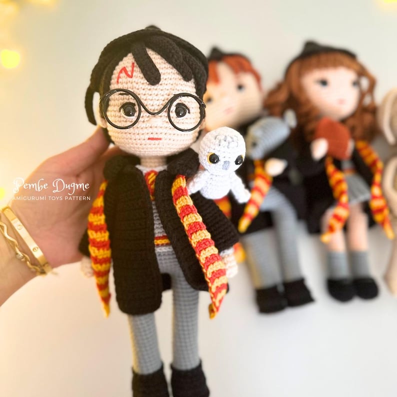 May include: Crocheted doll of a character with black hair, round glasses, and a lightning bolt scar. The doll wears a black robe, gray pants, and a red and gold scarf. A small white owl sits on the doll's shoulder. Other dolls are in the background.