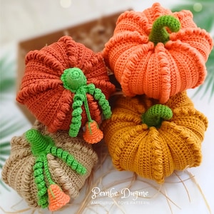 Peut inclure: Trois citrouilles en crochet orange, marron et beige avec des tiges vertes et des fleurs orange. Les citrouilles sont empilées les unes sur les autres. Le texte "Pembe Dugme AMIGURUMI TOYS PATTERN" est en bas de l'image.
