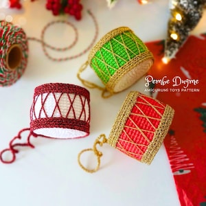May include: Three crocheted drum ornaments in red, green, and white with gold trim. The drums have a crisscross design and small gold loops. The text "Pembe Dugme Amigurumi Toys Pattern" is visible.
