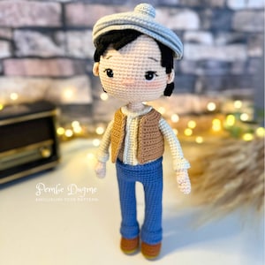 May include: A crocheted amigurumi doll wearing a blue shirt, brown vest, blue jeans, and a gray beret. The doll has black hair and a light brown complexion. The doll is standing on a white surface. The text "Pembe Dugme AMIGURUMI TOYS PATTERN" is visible below the doll.