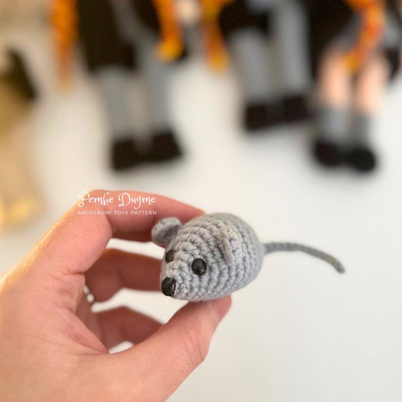 May include: A small, gray crocheted mouse toy with black bead eyes and a long tail. The mouse has small, rounded ears and a black nose. The text "Pembe Dugme Amigurumi Toys Pattern" is visible.