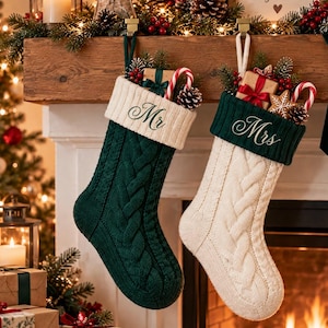 May include: Two cable-knit Christmas stockings hanging from a wooden mantel. The left stocking is dark green with a cream cuff embroidered with "Mr." The right stocking is cream with a dark green cuff embroidered with "Mrs." Both contain gifts.