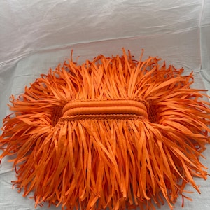 May include: A bright orange handbag featuring a dense, shaggy fringe made of long, thin strips of fabric. The bag has a central, horizontally oriented handle constructed from a braided or wrapped material, surrounded by the voluminous orange fringe.