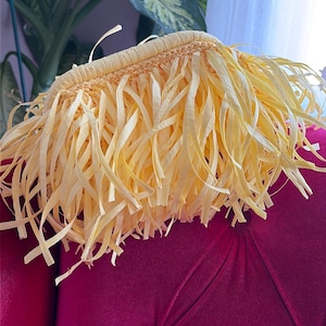 May include: A straw hat with a fringed brim. The hat is made of woven straw and has a light yellow color. The hat is sitting on a magenta velvet chair.