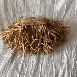 May include: A woven raffia clutch with long, fringed edges. The clutch is a natural tan color, with a rectangular body and a woven texture. The fringe hangs down from the bottom edge, creating a layered effect. The background is a white, textured fabric.