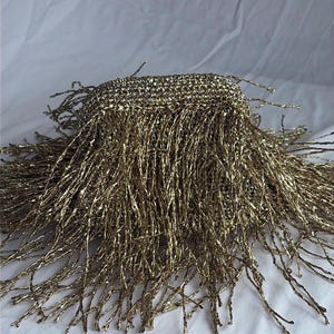 May include: A close-up of a gold-colored fringe trim. The trim has a woven top and long, twisted strands that hang down. The fringe is on a white background.