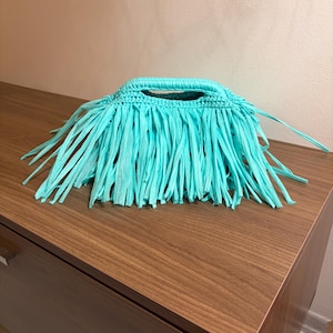 May include: A bright turquoise handbag crafted from crocheted material, featuring a structured top handle and a dense layer of long, thin fringe strips that hang down from the main body of the bag, creating a textured and layered appearance.