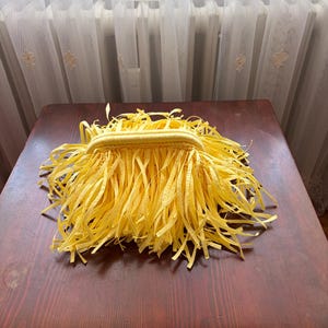 May include: A yellow clutch purse with a fringe design. The purse is made of a woven material and has long, thin strips of yellow fabric hanging from the top and sides. The purse is sitting on a dark brown wooden table.
