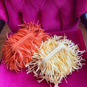 May include: Two fringed clutch bags, one orange and one cream, rest on a magenta velvet chair. The bags have woven handles and are made of long, thin strips of material, creating a textured, playful look. The chair has a tufted back.