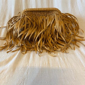 May include: A woven straw handbag with long, fringed edges. The bag is rectangular with a woven top and a fringe that hangs down. The color is a light tan, and the bag appears to be handmade.