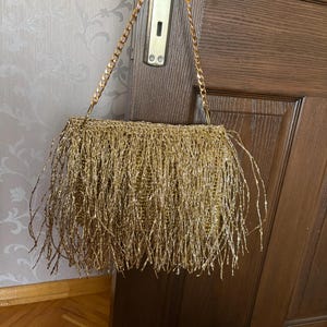 May include: A rectangular gold-colored handbag with a chain strap. The bag is covered in long, dangling strands, creating a fringed effect. The chain strap is also gold, and the bag hangs on a wooden door.