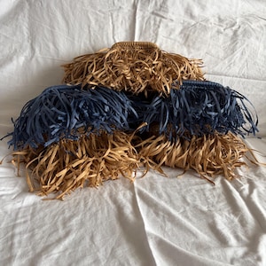 May include: Three straw hats with fringed edges. Two hats are natural straw color, and one is navy blue. The hats are arranged on a white sheet.