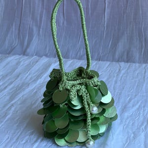 Può includere: Una piccola borsa a secchiello all'uncinetto color verde chiaro con un fitto strato di paillettes circolari sovrapposte di colore verde metallizzato. La borsa presenta un manico all'uncinetto e una chiusura a coulisse con due perle bianche.