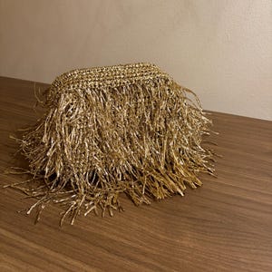 May include: A gold-colored, rectangular woven basket with long, dangling fringe. The fringe is made of shiny, metallic threads, creating a glamorous and textured appearance. The basket sits on a wooden surface.