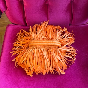 May include: An orange raffia clutch with a woven center section and long fringe. The clutch is set against a vibrant pink velvet chair, creating a striking contrast in color and texture. The bag's design suggests a bohemian or resort style.