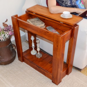 Może przedstawiać: Drewniany stolik boczny z ciepłym, czerwonobrązowym wykończeniem. Posiada górną powierzchnię, dolną półkę i szufladę. Stół zawiera pilota, książki i dekoracyjne świeczniki. Na górze stoi filiżanka herbaty.