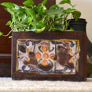 May include: A wooden planter box with a carved, painted design, filled with green and yellow leafy plants. The planter has a dark brown frame with a decorative panel featuring floral and scroll motifs in yellow, blue, and white.