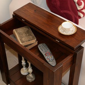 Könnte beinhalten: Ein dunkelbrauner Beistelltisch aus Holz mit einem hochklappbaren Deckel, der ein Staufach freilegt. Im Inneren befinden sich Bücher und eine Fernbedienung. Eine Teetasse und Untertasse stehen oben. Der Tisch hat ein unteres Regal mit dekorativen Kerzenhaltern.