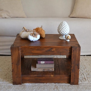Può includere: Un tavolino quadrato in legno tinto scuro con un ripiano inferiore con libri. Il tavolo è sormontato da zucche decorative e una statuina di ananas in ceramica bianca. Il tavolo è posto su un tappeto color crema.