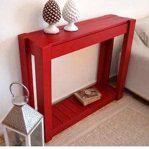 Può includere: Una console rosso vivo dal design rettangolare. Il tavolo presenta un ripiano inferiore ed è adornato da due ornamenti decorativi a forma di pigna. Un piccolo libro è appoggiato sul ripiano. A sinistra è visibile una lanterna argentata.