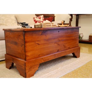 Puede incluir: Un baúl de almacenamiento rectangular de madera con un acabado marrón cálido y veta de madera visible. El baúl presenta una parte superior plana, un panel frontal simple con una pequeña cerradura y cuatro patas robustas con recortes.