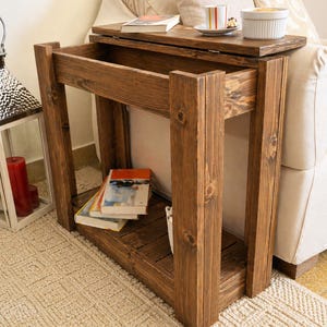 Puede incluir: Mesa auxiliar rústica de madera con tapa extraíble. Tiene un estante inferior para libros y una superficie superior con una taza, un platillo y un plato pequeño. La madera tiene un acabado marrón cálido. Mide aproximadamente 60 cm de alto.