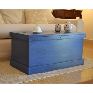 Pode incluir: Um grande baú de madeira azul com formato retangular. O baú é pintado em uma cor azul sólida e tem um tampo plano. Dois objetos decorativos de cerâmica em forma de ovo e uma tigela de cerâmica branca com juncos estão em cima do baú. O baú está sobre um tapete bege.