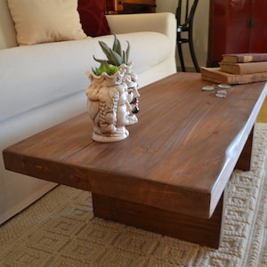 Op de afbeelding: Een rechthoekige, donkerbruine houten salontafel met een dik blad en een massieve voet. Een decoratieve keramische vaas met drie gezichten en een plant staat op de tafel, samen met een stapel boeken en wat munten. De tafel staat op een patroonkleed.