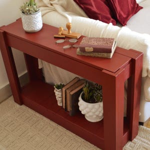 Può includere: Un tavolo console rettangolare in legno rosso scuro con un ripiano inferiore. La superficie superiore ospita un vaso bianco testurizzato con una pianta grassa, due timbri in legno, un paio di occhiali con montatura sottile e due libri.