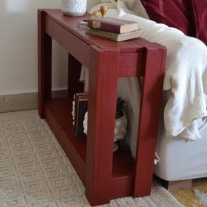 Può includere: Un tavolo console stretto e rettangolare con una finitura rossa opaca, dotato di una superficie superiore piatta e di un ripiano inferiore. Il tavolo è costruito con spesse gambe in legno verticali e travi di supporto orizzontali, offrendo una struttura robusta.