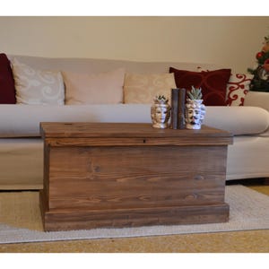 Peut inclure: Un grand coffre en bois rectangulaire avec un couvercle à charnière, teinté d'une teinte marron chaude. Le coffre est placé devant un canapé crème avec des coussins décoratifs. Deux vases décoratifs en forme de tête et un livre sont posés dessus.