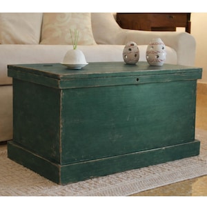 Può includere: Un baule rettangolare in legno con una finitura verde scuro usurata. Il baule presenta un coperchio piatto con un piccolo foro circolare per la chiave. Sopra il baule sono posizionati tre oggetti decorativi in ceramica bianca, tra cui una cupola.