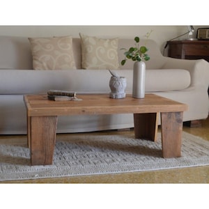 Peut inclure: Une table basse rectangulaire en bois avec une finition naturelle. La table est soutenue par quatre pieds épais. Un petit tapis est sous la table. Un vase avec de la verdure, un mortier et un pilon, et une petite brosse sont sur la table. La table mesure environ 120 cm de long.