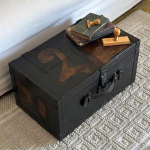 Pode incluir: Um baú de armazenamento retangular preto com uma alça de metal escuro e um padrão de redemoinho marrom na tampa. Dois livros antigos com capas gastas e dois carimbos de madeira com cabos claros estão colocados em cima do baú.