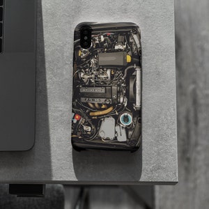 May include: A black phone case with a detailed image of a car engine. The engine is made up of various metal parts, including hoses, wires, and a black air filter.