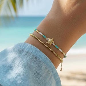 May include: Two gold beaded bracelets. One bracelet features a small gold turtle charm and turquoise, white, and gold beads. The other bracelet is a simple gold beaded design. The bracelets are worn on a wrist, with a blue shirt in the background.